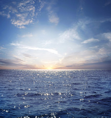 Rising sun on the horizon above a calm ocean or sea. On the blue sky white clouds