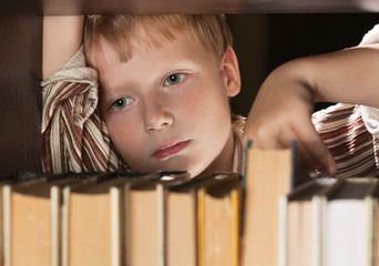 The boy at a bookcase gets the book. The boy look tired.