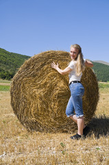 Naklejka na ściany i meble Blonde girl leaning against a rolling haystack.