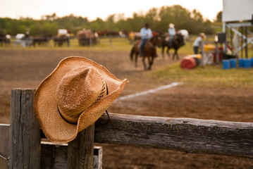 Cowboy hat hung on the fence at a rodeo with horses and riders - obrazy, fototapety, plakaty
