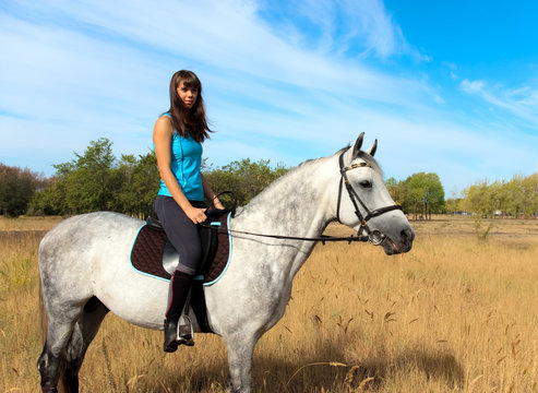 A young woman horseback rider in a blue top sits on a dapple-grey horse in a sunny field of dry grass under a blue sky.