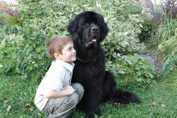 Мальчик в обнимку с черным ньюфаундлендом на фоне зелени