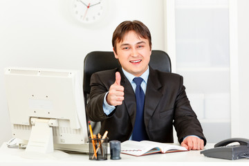 Business man sitting at desk and showing thumbs up gesture.