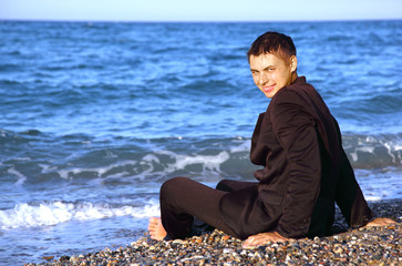 Barefooted smiling man in suit sits on stone coast at evening