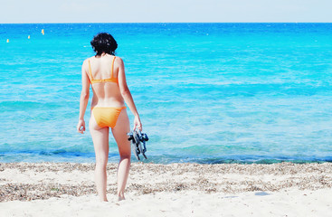 Woman in orange bikini holding flops on the shoreline