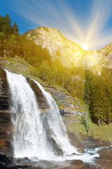 Alpine waterfall in mountain forest in sun rays under blue sky. - 29419774