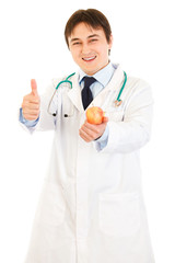 Smiling  doctor holding apple and showing thumbs up gesture
