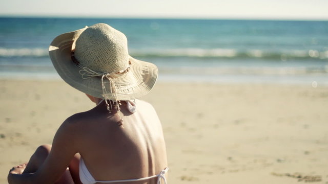 Woman in hat sitting on the beach looking out to the sea
