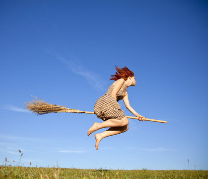 Young red-haired witch on broom flying over green grass field