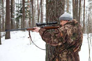 Hunter in camouflage aiming with sniper rifle at winter forest.