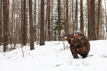 Hunter in camouflage aiming with sniper rifle at winter forest.
