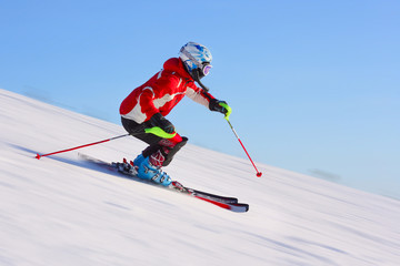 Skier in red jacket riding on hill. Motion blurred snow and clear blue sky