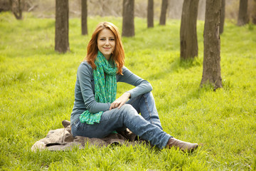 Beautiful red-haired girl sitting at green grass at park.