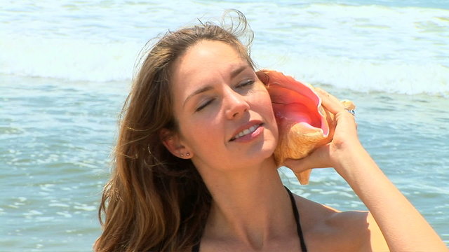 Young woman in bikini listening to conch shell by ocean surf