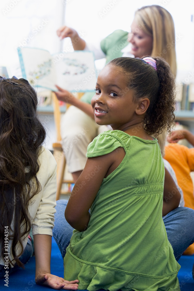 Wall mural Kindergarten teacher reading to children in library, girl lookin