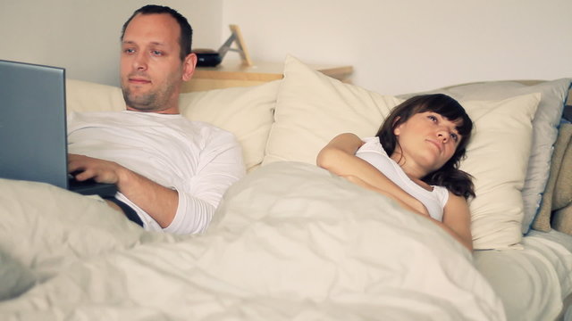 Unhappy woman in bed and a man working on computer