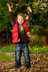 Happy red haired boy playing with autumn leaves in park