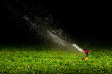 lawn sprinkler spraying water over green grass at night - obrazy, fototapety, plakaty