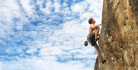 A young man climbs on a cliff over blue sky background