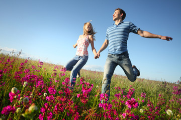 Young happy man and woman running on meadow with fluffy dandelions on blue sky background
