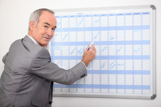 Older Businessman Writing On A Wall Planner