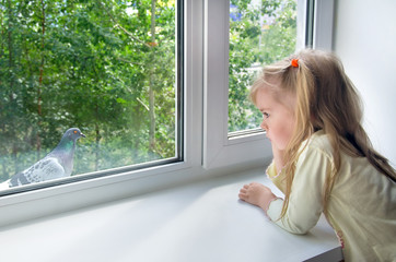 A sad little girl looks at a pigeon outside the window