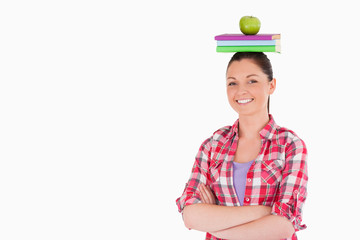 Pretty female holding an apple and books on her head while stand