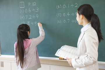 黒板の問題を解く小学生女子と横に立つ女性教師