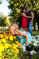 Fototapeta premium Little girl smelling flowers, behind her are happy parents.