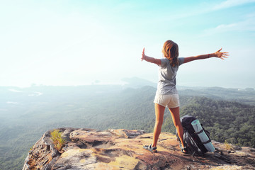 Young woman standing with raised hands with a backpack on the cliff's edge and looking into a wide valley