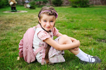 Young school girl with pink bagpack sits on grass