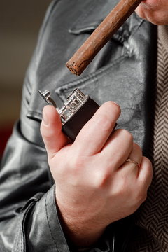 Мужчина в кожаной куртке раскуривает толстую сигару дорогой зажигалкой