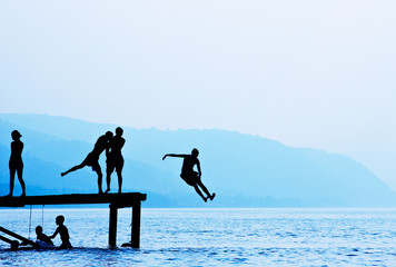 Silhouettes of kids who jump off dock on the lake - obrazy, fototapety, plakaty