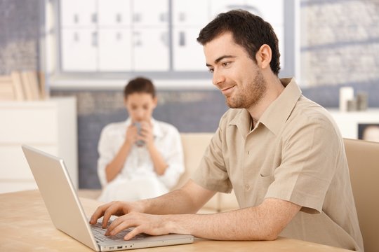 Young man using laptop at home woman in background