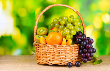 Ripe juicy fruits in basket on wooden table on green background