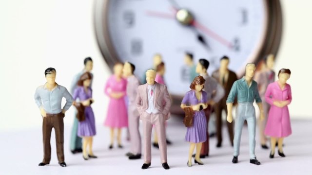 Group of little toy men and women stand in front of big clock