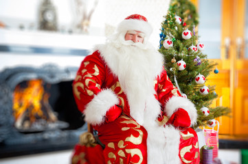 Santa sitting at the Christmas tree, near fireplace and looking