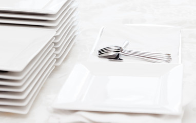 Stack of square white plates and silver spoons on table