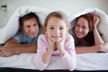 Obraz na płótnie Canvas Cheerful girl under bed cover with parents