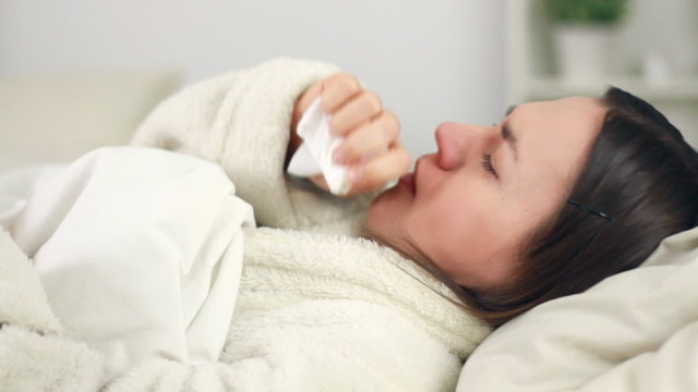 Sick woman in bed coughing and blowing nose in paper tissue