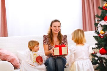 Two twins girl presenting gift to mother near Christmas tree