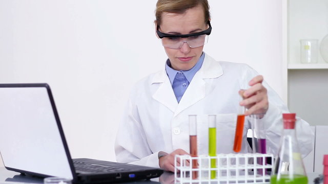 Female scientist examines test tubes and writing on laptop