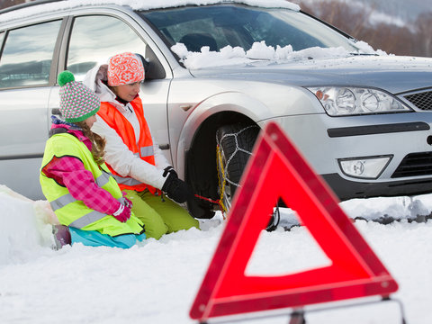 Winter, Woman With Child Putting Snow Chains Onto Tyre Of Car