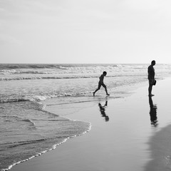 small boy plays by the ocean as his dad watches on