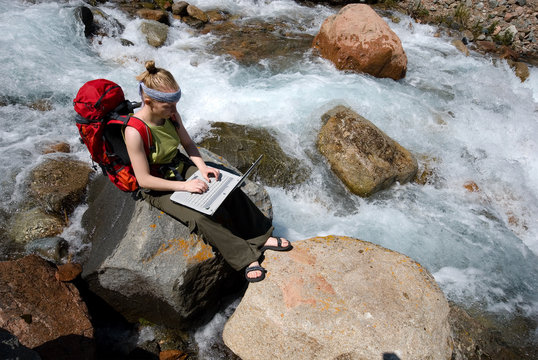 Young girl sits on the mountain river and works on a computer