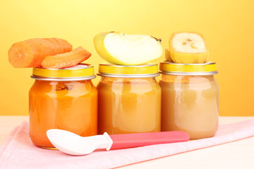 Jars of baby puree with spoon on napkin on yellow background