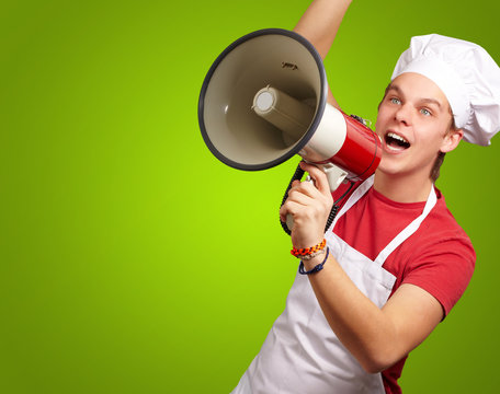 portrait of young cook man shouting with megaphone over green ba