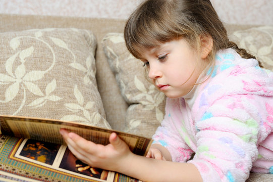 The little girl reads the book lying on a sofa