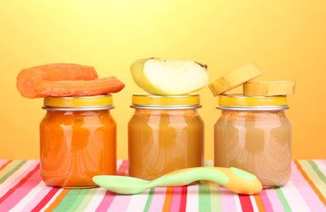 Jars of baby puree with spoon on napkin on yellow background