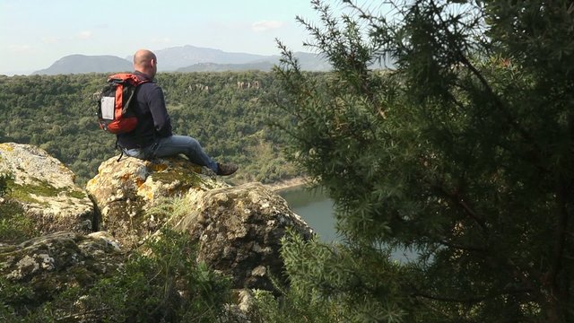 Adult man with binoculars sitting on rock, looking at lake